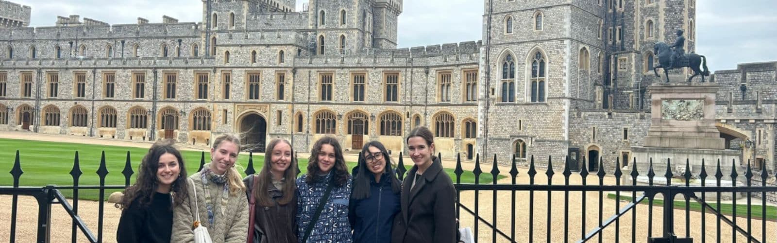 Six students stand outside of a metal gate leading to a massive stone castle. 